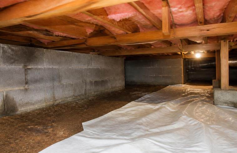 Interior of a crawl space with a dirt floor partly covered by loose white plastic sheeting, concrete block walls, and insulated floor joists above